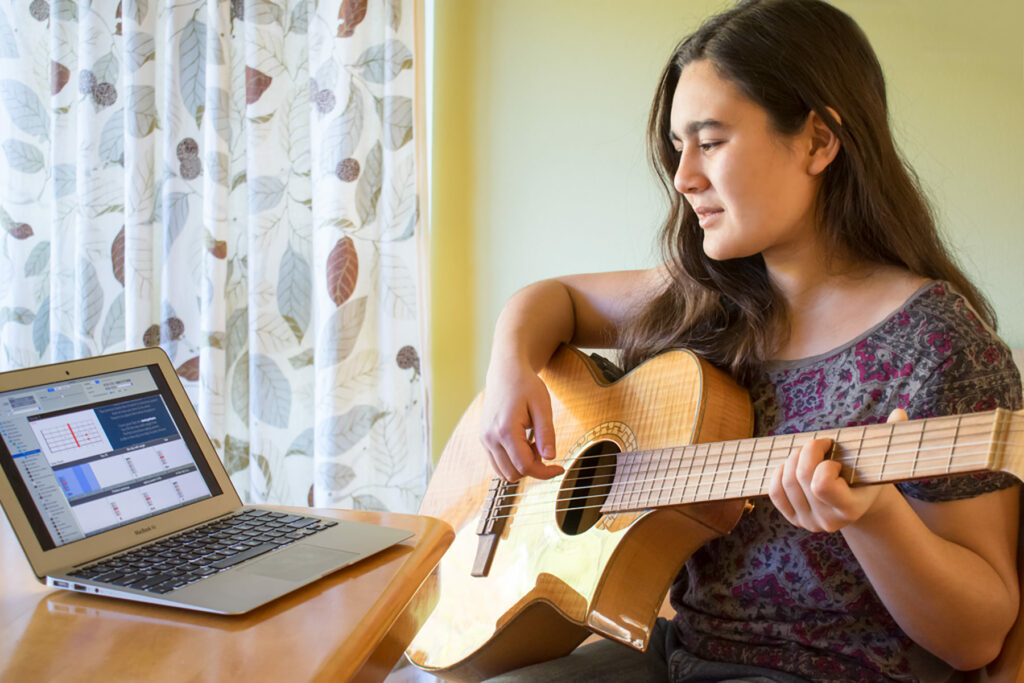 Young woman playing guitar in front of laptop.